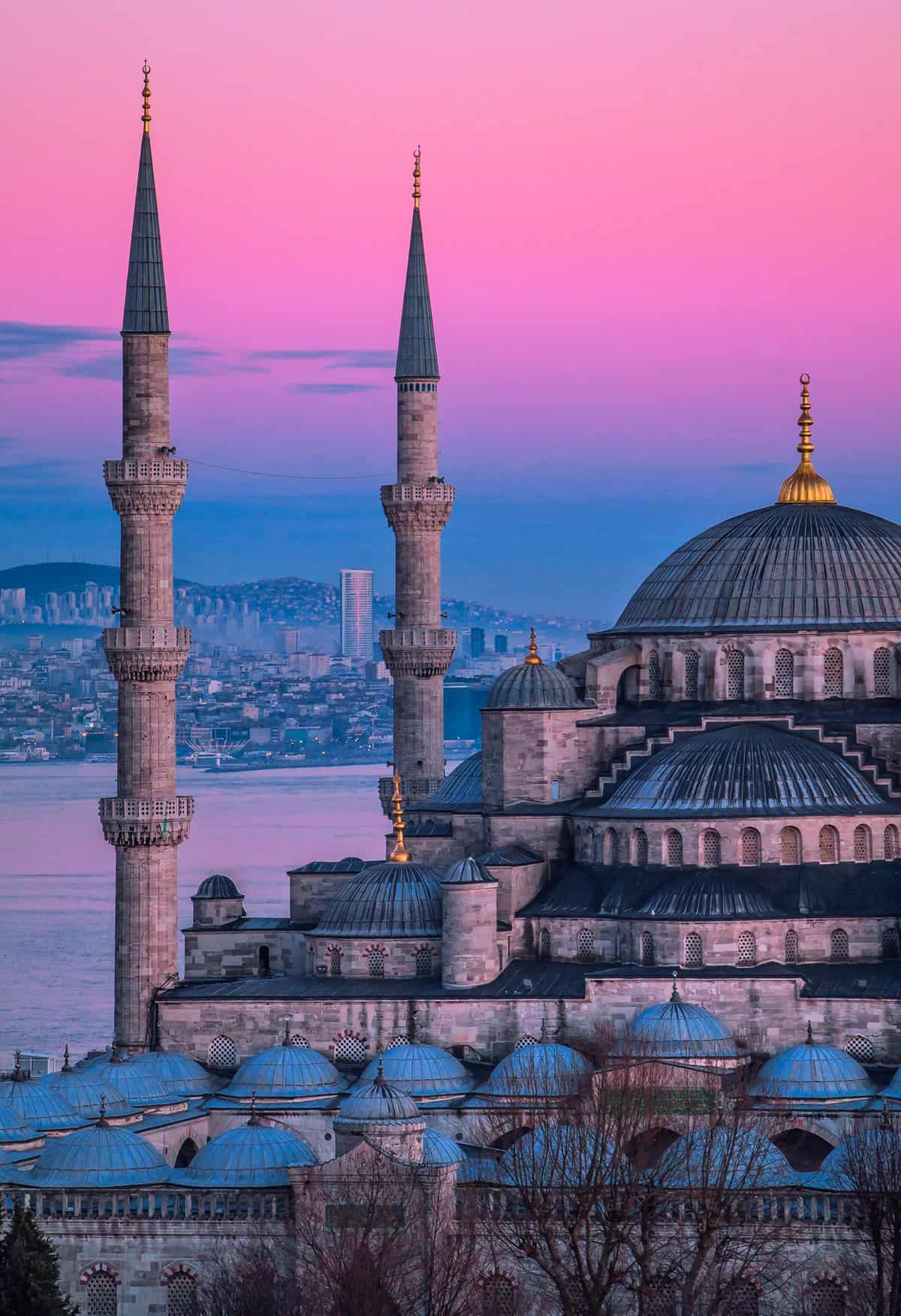 An architectural view of the Blue Mosque (Sultan Ahmed Mosque) in Istanbul, with its prominent minarets and domes, against a pink and purple sky at dusk, overlooking city buildings and a body of water—a perfect sight for those on an Explora Journeys luxury travel experience.